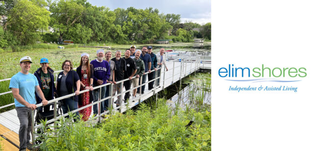 A group of smiling residents and staff from Elim Shores, an independent and assisted living community, stands on a dock next to a serene waterway surrounded by lush greenery. The group, dressed in casual attire, celebrates the new outdoor amenities, including a dock and pontoon boat, made possible by generous donations.