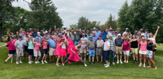 A large group of people posing playfully at a golf event, smiling and raising their hands in the air. They are dressed in a variety of casual and sporty outfits, with some in pink and colorful attire. The group, ranging in age, is enjoying a fun and silly moment on the golf course, surrounded by lush greenery and trees.