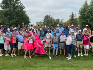 A large group of people posing playfully at a golf event, smiling and raising their hands in the air. They are dressed in a variety of casual and sporty outfits, with some in pink and colorful attire. The group, ranging in age, is enjoying a fun and silly moment on the golf course, surrounded by lush greenery and trees.