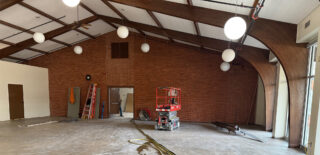 A spacious chapel under renovation with a brick wall, exposed ceiling beams, and construction equipment scattered throughout.